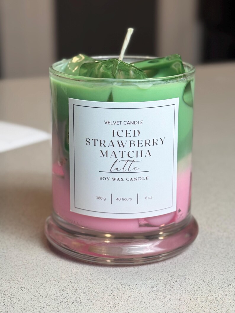 May include: A soy wax candle in a clear glass jar with a white label reading VELVET CANDLE ICED STRAWBERRY MATCHA latte SOY WAX CANDLE 180 g | 40 hours | 8 oz. The candle features layers of pink and green wax with green wax ice cubes.
