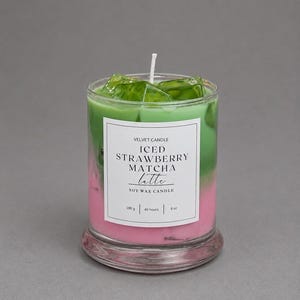 May include: A soy wax candle in a clear glass jar featuring a layered pink and green design topped with green wax ice cubes. The white label reads VELVET CANDLE ICED STRAWBERRY MATCHA latte SOY WAX CANDLE 180 g | 40 hours | 8 oz with a central wick.