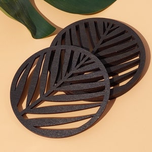 May include: Two black wooden coasters featuring a palm leaf design. The coasters are round and have a layered appearance.