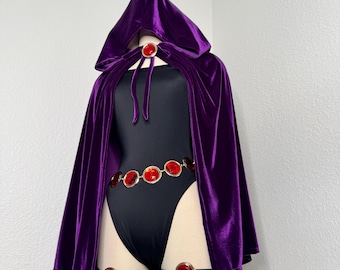 May include: A purple velvet hooded cloak and black bodysuit costume. The costume includes a belt and leg bands with red jewel accents. The cloak is fastened with a red jewel clasp.