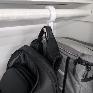 Puede incluir: Un gancho de plástico blanco cuelga de una barra de armario blanca, sosteniendo dos mochilas. Una es negra, la otra gris con detalles negros. La barra es de madera pintada de blanco.