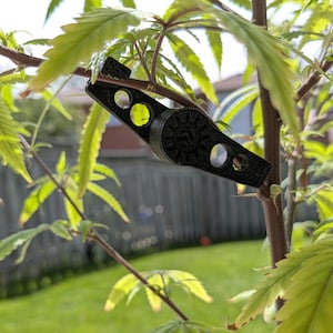 Puede incluir: Una herramienta negra de entrenamiento de plantas, impresa en 3D, está sujeta a una rama de planta de cannabis. La herramienta tiene una serie de agujeros circulares y un diseño decorativo en forma de engranaje. El fondo muestra follaje verde y una escena exterior borrosa.