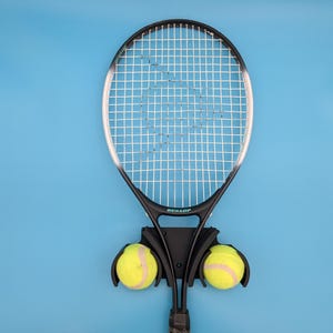 May include: A black tennis racket with a white string pattern and the word "Dunlop" printed on the handle. The racket is mounted on a black wall rack with two yellow tennis balls.