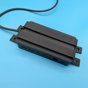 Puede incluir: Un dispositivo electrónico rectangular negro con una serie de pequeñas aberturas rectangulares a un lado. El dispositivo tiene una parte superior acanalada y está conectado a un cable negro. El fondo es azul claro liso.