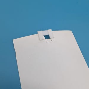 Op de afbeelding: Een wit rechthoekig papier met een witte plastic clip bevestigd aan de bovenkant. Het papier heeft een gescheurde rand. De achtergrond is effen lichtblauw.
