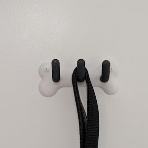 May include: A white, bone-shaped wall hook with three black hooks. A black dog lead hangs from the central hook. The hook is mounted on a white wall.