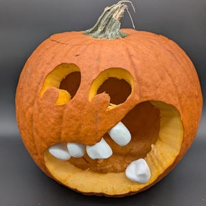 Könnte beinhalten: Ein geschnitzter orangefarbener Kürbis mit großen Augen- und Mundöffnungen. Der Kürbis hat einen grünen Stiel und weiße Zähne. Der Kürbis ist eine Halloween-Dekoration.