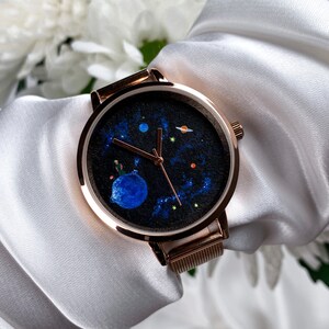 May include: A rose gold watch with a black face featuring a painted design of a blue planet, stars, and a small figure. The watch has a mesh band.