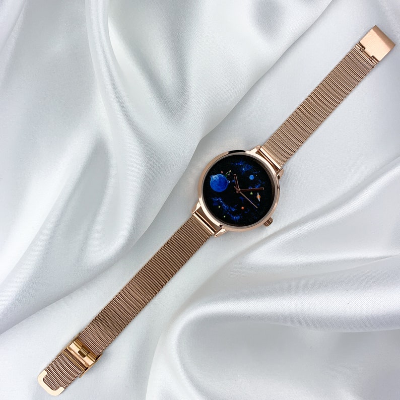May include: A rose gold mesh band watch with a round face featuring a blue and black galaxy design.