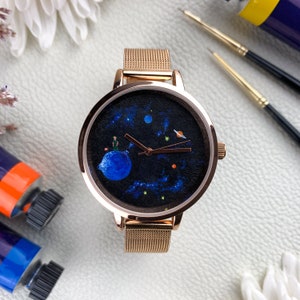 May include: A rose gold watch with a black face featuring a painted design of a planet with a small figure on it, surrounded by stars and other planets. The watch has a mesh band.