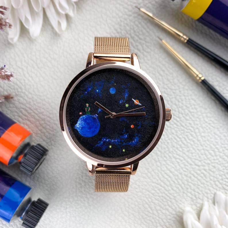 May include: A rose gold watch with a black face featuring a painted design of a planet with a small figure on it, surrounded by stars and other planets. The watch has a mesh band.