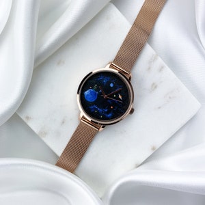 May include: A rose gold mesh band watch with a black face featuring a painted design of a blue planet and stars.