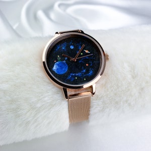 May include: A rose gold watch with a black face featuring a painted design of the Little Prince on a planet. The watch has a mesh band.
