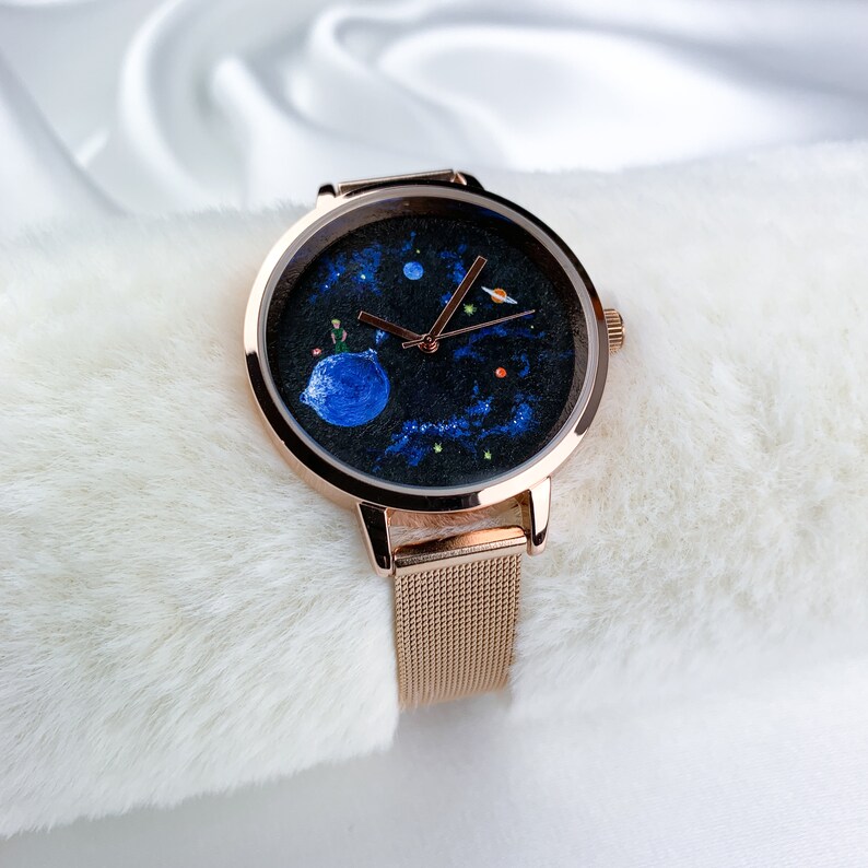 May include: A rose gold watch with a black face featuring a painted design of the Little Prince on a planet. The watch has a mesh band.