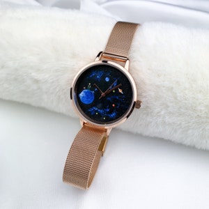 May include: A rose gold watch with a mesh band and a round face. The face has a black background with a blue planet, stars, and a small figure on the planet.