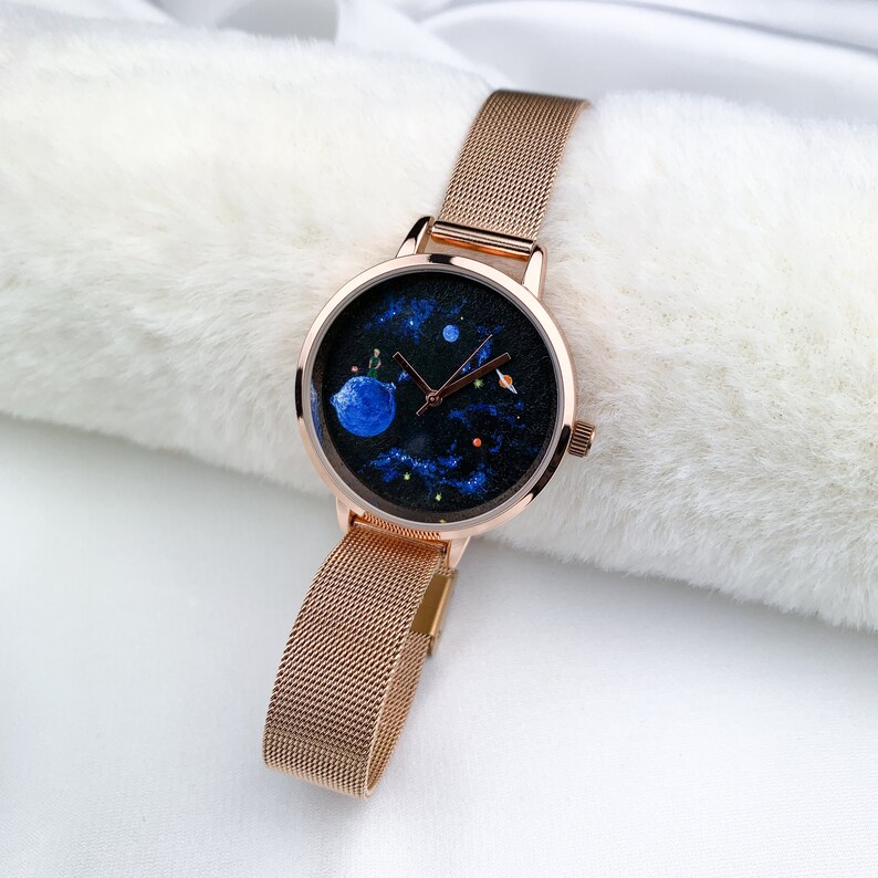 May include: A rose gold watch with a mesh band and a round face. The face has a black background with a blue planet, stars, and a small figure on the planet.