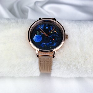 May include: A rose gold watch with a black face featuring a painted design of The Little Prince on his asteroid. The watch has a mesh band.