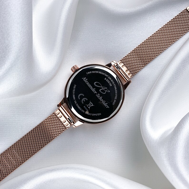 May include: The back of a rose gold watch with a black face. The watch face has the text "Alexander Schr&ouml;der" and "8 bar water resistant" printed on it.