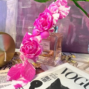 May include: A floral headpiece with pink and white flowers and leaves, placed on a perfume bottle. The bottle is next to a Vogue magazine with a black cat illustration. The background features a painting with green and pink hues.