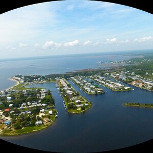 Puede incluir: Vista aérea de una comunidad costera con casas, muelles y barcos. La imagen muestra una península con una vía fluvial y una masa de agua bajo un cielo azul con nubes. La escena es un día soleado.