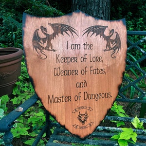 May include: A wooden shield with a burnt finish features two dragons flanking a text inscription that reads "I am the Keeper of Lore, Weaver of Fates, and Master of Dungeons."