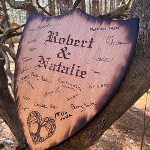 May include: Wooden shield-shaped sign with the names "Robert & Natalie" in elegant script. The sign is adorned with signatures and a heart-shaped tree design. The edges are dark, and the wood has a natural grain.
