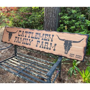 May include: A rustic wooden sign with the text "Cattlemen Family Farm" and a skull with horns on each end. The sign is on a black metal bench and the text "Est. 2021" is on the bottom right corner.