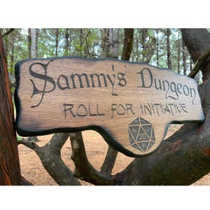 May include: A wooden sign with a dark brown stain and black edging. The sign reads "Sammy's Dungeon Roll for Initiative" with a 20-sided die graphic below the text.