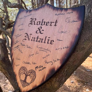 May include: A wooden shield-shaped sign with the names "Robert & Natalie" in elegant script. The sign is adorned with signatures and a heart-shaped tree design. The edges are dark, and the wood has a natural grain.