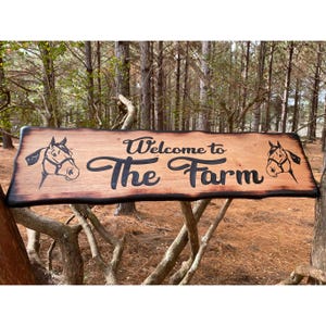 May include: A wooden sign with a burnt edge, featuring the text "Welcome to The Farm" in black lettering. Two horse head silhouettes are on either side of the text.