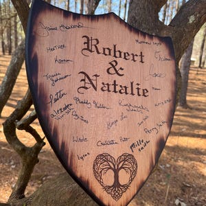 May include: A wooden shield-shaped sign with the names "Robert & Natalie" in elegant script. The sign is adorned with signatures and a heart-shaped tree design. The edges are dark, and the wood has a natural grain.