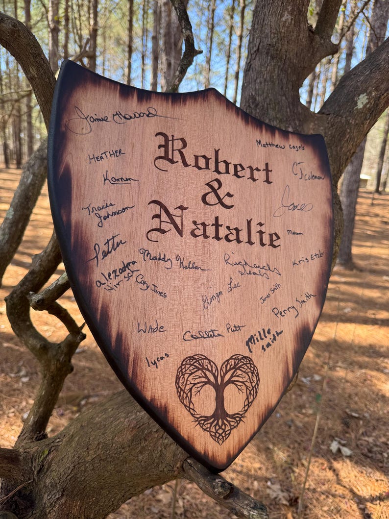 May include: A wooden shield-shaped sign with the names "Robert & Natalie" in elegant script. The sign is adorned with signatures and a heart-shaped tree design. The edges are dark, and the wood has a natural grain.