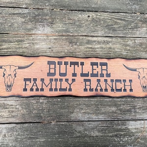 May include: A wooden sign with a rustic, burnt finish. The sign reads "BUTLER FAMILY RANCH" in black lettering. Two stylized bull skulls are on either side of the text.