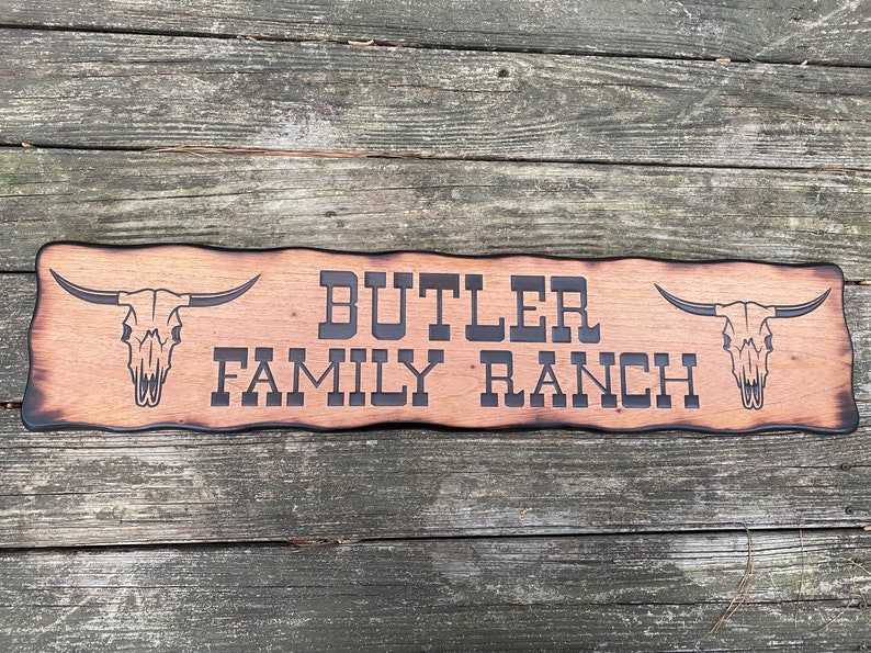 May include: A wooden sign with a rustic, burnt finish. The sign reads "BUTLER FAMILY RANCH" in black lettering. Two stylized bull skulls are on either side of the text.