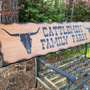 May include: A wooden sign with a burnt edge, featuring a black silhouette of a longhorn skull and the text "Cattlemen Family Farm" in black lettering.