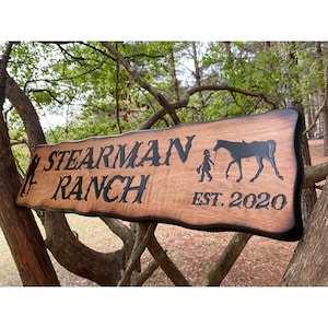 May include: A rustic wooden sign with the words "Stearman Ranch Est. 2020" burned into the wood. The sign also features a silhouette of a horse and rider.