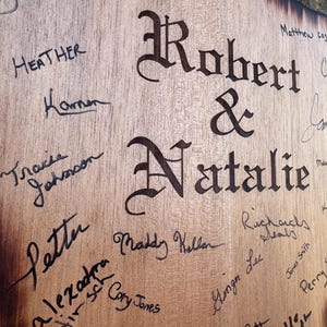 May include: A wooden sign with the names "Robert & Natalie" in large, ornate lettering. The sign is covered in signatures in black ink, with names like "Jaime Haward" and "Heather" visible. The wood has a natural grain.