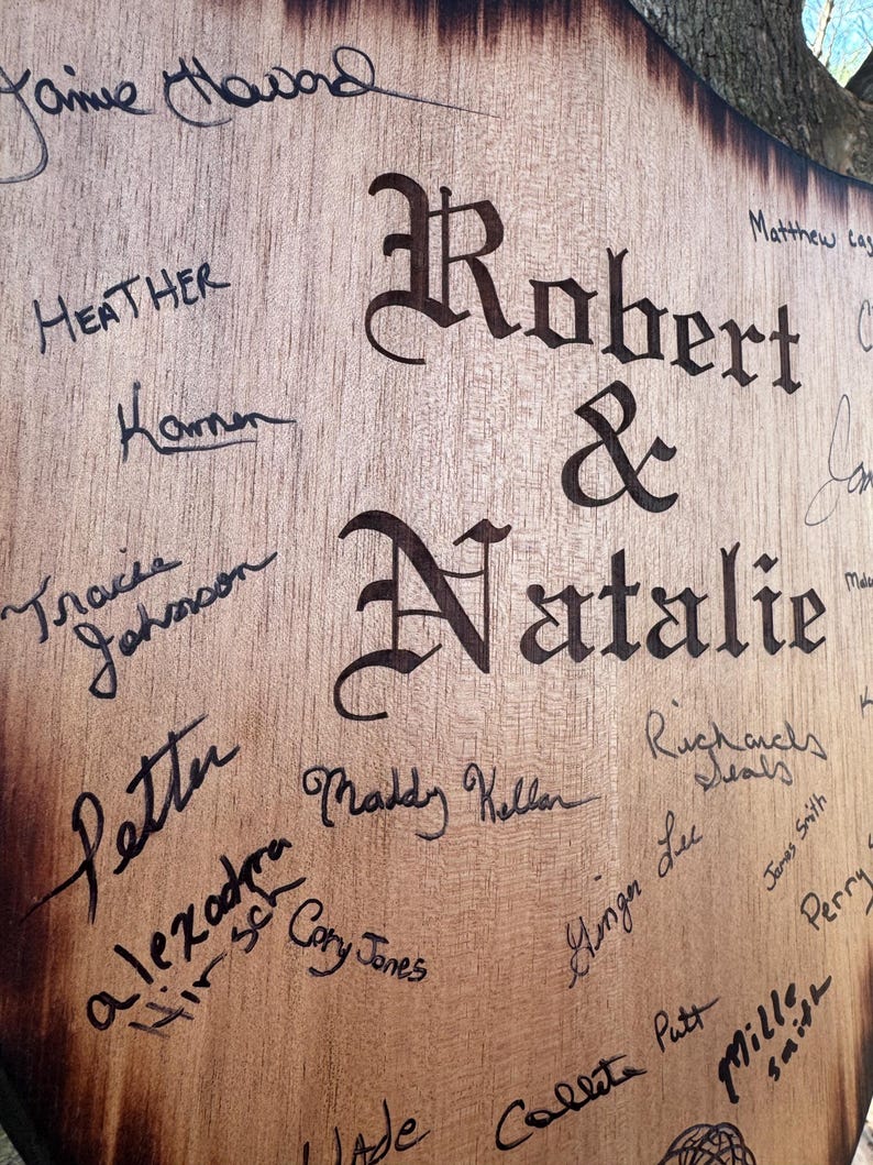 May include: A wooden sign with the names "Robert & Natalie" in large, ornate lettering. The sign is covered in signatures in black ink, with names like "Jaime Haward" and "Heather" visible. The wood has a natural grain.
