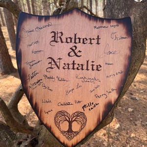 May include: Wooden shield-shaped sign with the names "Robert & Natalie" in elegant script. The sign is adorned with signatures and a heart-shaped tree design, perfect for a wedding or anniversary. The edges are dark brown.