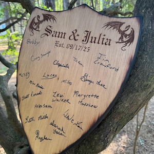 May include: A shield-shaped wooden plaque with burnt edges, featuring two engraved dragons and the text "Sam & Julia" above "Est. 09/17/25". The surface is covered in various handwritten signatures in black ink, creating a guest book style display.