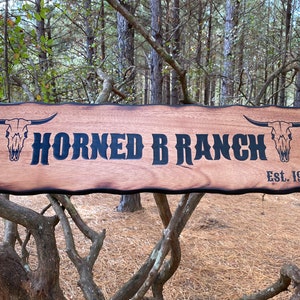 May include: A rustic wooden sign with a burnt edge, featuring the text "Horned B Ranch Est. 1998" in black lettering. Two stylized bull skulls are on either side of the text.