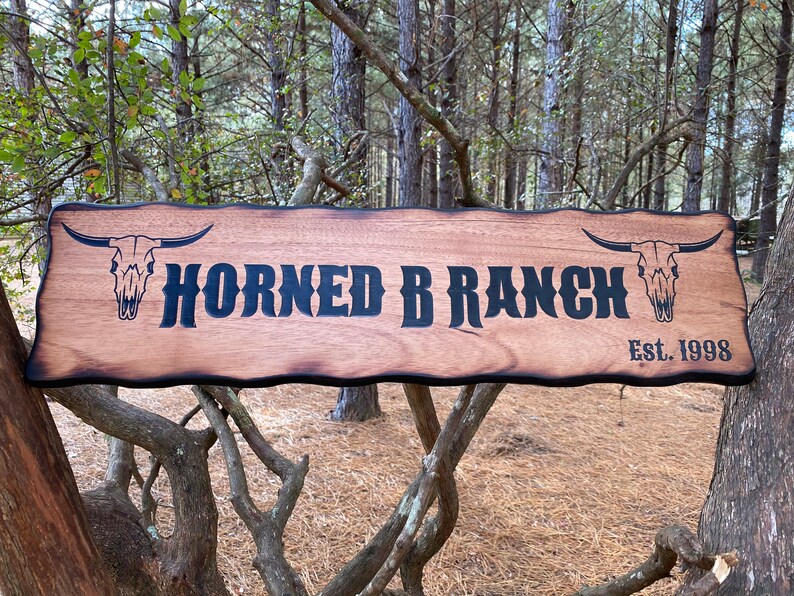 May include: A rustic wooden sign with a burnt edge, featuring the text "Horned B Ranch Est. 1998" in black lettering. Two stylized bull skulls are on either side of the text.