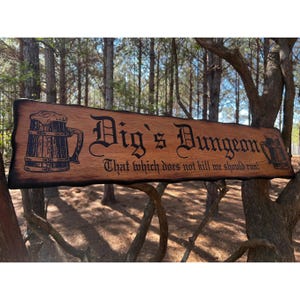 May include: A wooden sign with a burnt finish reads "Dig's Dungeon" in a black gothic font. The sign is decorated with two images of beer steins and the quote "That which does not kill me should run!"