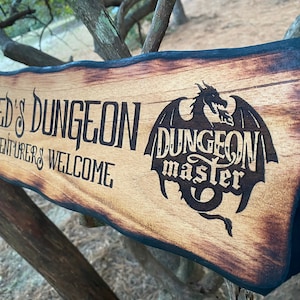 以下が含まれることがあります： 焦げ付いた縁のある木製の看板で、「SYED'S DUNGEON ADVENTURERS WELCOME」という文字と、「DUNGEON master」という文字の入った黒いシルエットのドラゴンが描かれている。