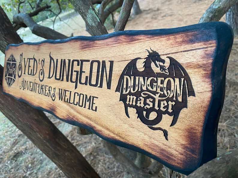 以下が含まれることがあります： 焦げ付いた縁のある木製の看板で、「SYED'S DUNGEON ADVENTURERS WELCOME」という文字と、「DUNGEON master」という文字の入った黒いシルエットのドラゴンが描かれている。