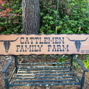 May include: A wooden sign with a burnt edge, featuring the text "Cattlemen Family Farm Est. 2021" in black lettering. The sign is decorated with two black silhouettes of steer skulls.