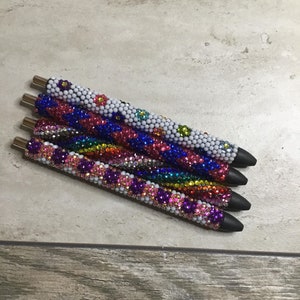 Pode incluir: Cinco canetas coloridas com strass e base branca. As canetas têm uma ponta de tinta preta e estão dispostas em uma pilha.