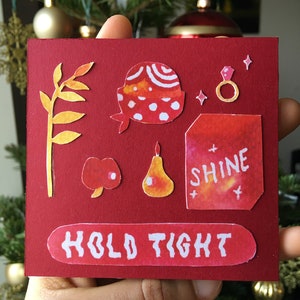 Puede incluir: Tarjeta roja con una variedad de recortes de papel e ilustraciones de acuarela. La tarjeta presenta una manzana roja, una pera, un anillo, una rama con hojas, un sombrero rojo y blanco y un cuadrado rojo con la palabra "shine" en texto blanco. La tarjeta también tiene las palabras "hold tight" en texto blanco sobre un fondo rojo.