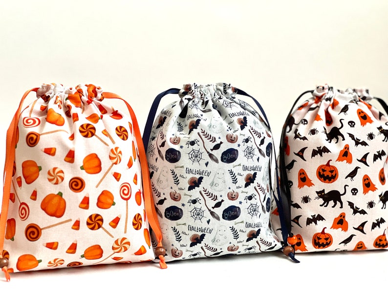 May include: Three Halloween-themed drawstring bags with different patterns. The first bag has a white background with orange pumpkins, candy canes, and candy corn. The second bag has a white background with black bats, pumpkins, ghosts, and the words "Boo!" and "Halloween". The third bag has a white background with black bats, pumpkins, ghosts, and black cats.