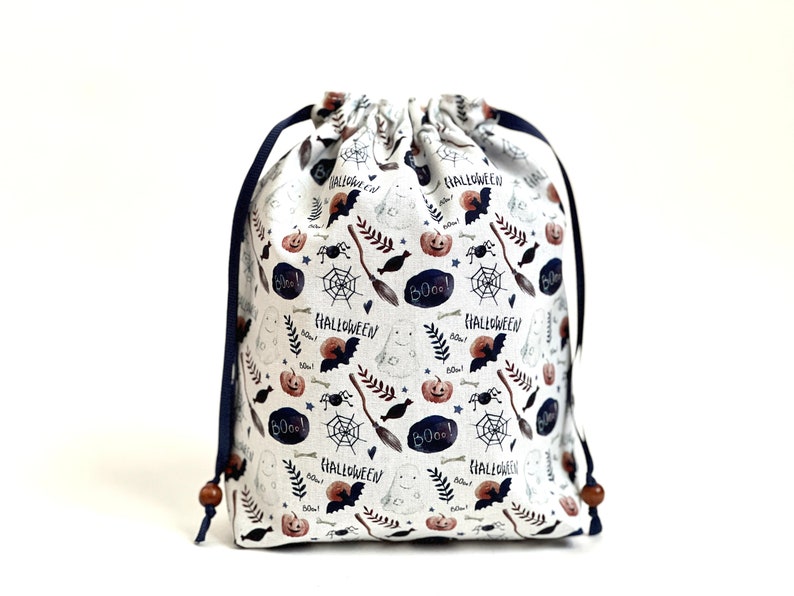 May include: White drawstring bag with a Halloween themed print. The print features bats, pumpkins, ghosts, spiders, bones, and the words "Boo!" and "Halloween".